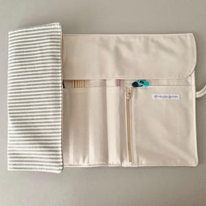 May include: Cream-coloured knitting needle roll with a grey and white striped flap. The roll has multiple pockets, a zip closure, and a white cord for carrying. A few knitting needles and stitch markers are visible.