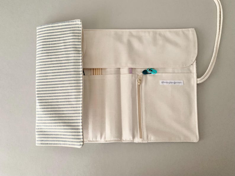 May include: Cream-coloured knitting needle roll with a grey and white striped flap. The roll has multiple pockets, a zip closure, and a white cord for carrying. A few knitting needles and stitch markers are visible.