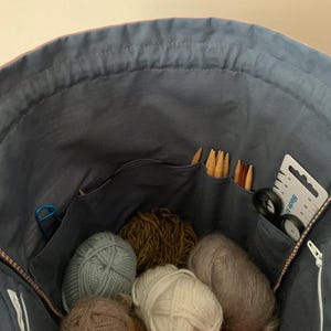 Pode incluir: Uma bolsa de tecido azul cheia de v&aacute;rios materiais de tric&ocirc;. V&aacute;rios novelos de l&atilde; em tons de cinza, marrom e branco s&atilde;o vis&iacute;veis. Agulhas de tric&ocirc;, tesouras e um medidor est&atilde;o nos bolsos, sugerindo um tema artesanal.