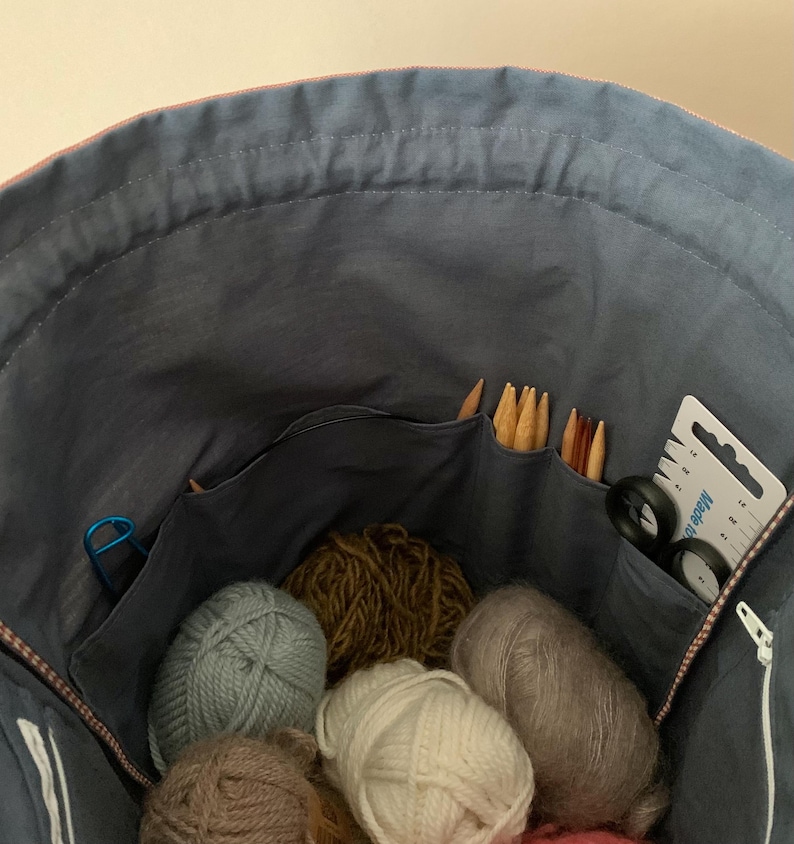Pode incluir: Uma bolsa de tecido azul cheia de v&aacute;rios materiais de tric&ocirc;. V&aacute;rios novelos de l&atilde; em tons de cinza, marrom e branco s&atilde;o vis&iacute;veis. Agulhas de tric&ocirc;, tesouras e um medidor est&atilde;o nos bolsos, sugerindo um tema artesanal.