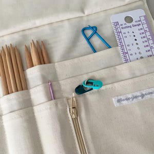 May include: Cream canvas knitting needle organiser with various knitting tools. Includes wooden needles, a crochet hook, stitch markers, a blue stitch holder, and a knitting gauge. The organiser has a gold zip and a label that reads "thereyougrows".