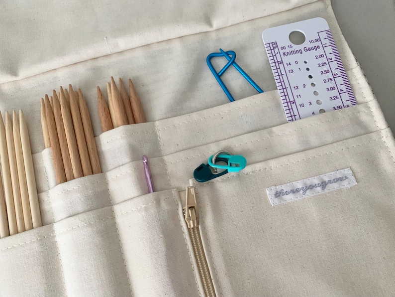 May include: Cream canvas knitting needle organiser with various knitting tools. Includes wooden needles, a crochet hook, stitch markers, a blue stitch holder, and a knitting gauge. The organiser has a gold zip and a label that reads "thereyougrows".