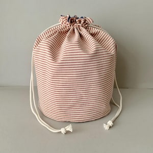 Pode incluir: Uma bolsa cil&iacute;ndrica com cord&atilde;o feita de tecido com listras horizontais vermelhas e brancas. A parte superior da bolsa &eacute; franzida e fechada com um cord&atilde;o branco. A bolsa foi projetada para guardar e transportar itens.