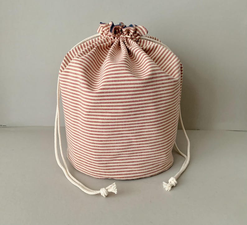 Pode incluir: Uma bolsa cil&iacute;ndrica com cord&atilde;o feita de tecido com listras horizontais vermelhas e brancas. A parte superior da bolsa &eacute; franzida e fechada com um cord&atilde;o branco. A bolsa foi projetada para guardar e transportar itens.