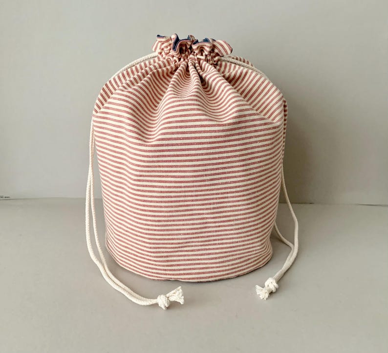 Pode incluir: Uma bolsa cil&iacute;ndrica com cord&atilde;o com um padr&atilde;o de listras vermelhas e brancas. A bolsa &eacute; feita de tecido e tem um fecho de cord&atilde;o branco. A bolsa tem aproximadamente 20 cm de di&acirc;metro e 25 cm de altura.