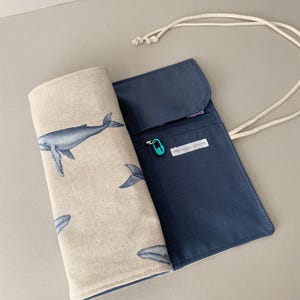 Puede incluir: Un estuche para l&aacute;pices azul y beige con tem&aacute;tica de ballenas. El estuche est&aacute; abierto, revelando un interior azul marino y un exterior beige estampado con ballenas azules. Se adjunta un cord&oacute;n blanco. Se ve un peque&ntilde;o imperdible turquesa.