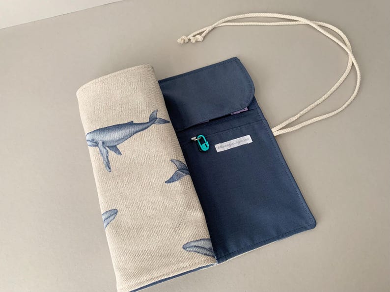 Puede incluir: Un estuche para l&aacute;pices azul y beige con tem&aacute;tica de ballenas. El estuche est&aacute; abierto, revelando un interior azul marino y un exterior beige estampado con ballenas azules. Se adjunta un cord&oacute;n blanco. Se ve un peque&ntilde;o imperdible turquesa.