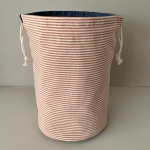 Pode incluir: Um saco de armazenamento cil&iacute;ndrico com um padr&atilde;o de listras vermelhas e brancas. A bolsa tem um interior azul e um fecho de cord&atilde;o com corda branca. A bolsa &eacute; feita de tecido e &eacute; adequada para v&aacute;rias necessidades de armazenamento.