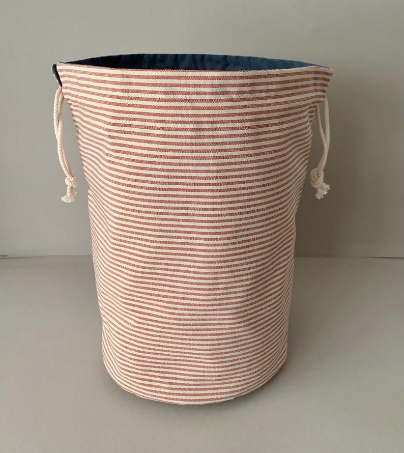 Pode incluir: Um saco de armazenamento cil&iacute;ndrico com um padr&atilde;o de listras vermelhas e brancas. A bolsa tem um interior azul e um fecho de cord&atilde;o com corda branca. A bolsa &eacute; feita de tecido e &eacute; adequada para v&aacute;rias necessidades de armazenamento.