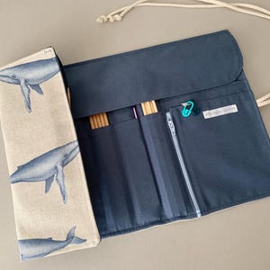Puede incluir: Estuche para agujas de tejer azul marino con solapa beige estampada con ilustraciones de ballenas azules. El estuche est&aacute; abierto, revelando bolsillos y contenido. Incluye un bolsillo con cremallera y ranuras para agujas de tejer y accesorios. Se adjunta un cord&oacute;n blanco.