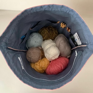 Pode incluir: Uma sacola azul marinho com acabamento rosa, cheia de bolas de l&atilde; de v&aacute;rias cores, agulhas de tric&ocirc; e outros materiais de artesanato. A sacola tem v&aacute;rios bolsos e um fecho de z&iacute;per.