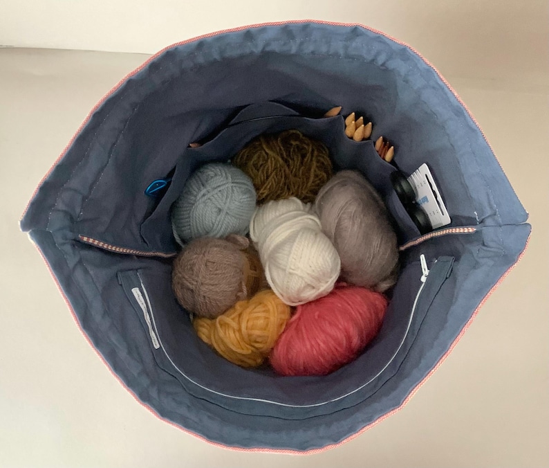 Pode incluir: Uma sacola azul marinho com acabamento rosa, cheia de bolas de l&atilde; de v&aacute;rias cores, agulhas de tric&ocirc; e outros materiais de artesanato. A sacola tem v&aacute;rios bolsos e um fecho de z&iacute;per.