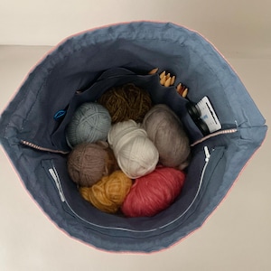Pode incluir: Uma bolsa de projeto de tric&ocirc; de tecido azul com acabamento rosa, cheia de bolas de l&atilde; de v&aacute;rias cores, agulhas de tric&ocirc; e pequenos acess&oacute;rios. A bolsa tem v&aacute;rios bolsos e um fecho de correr.