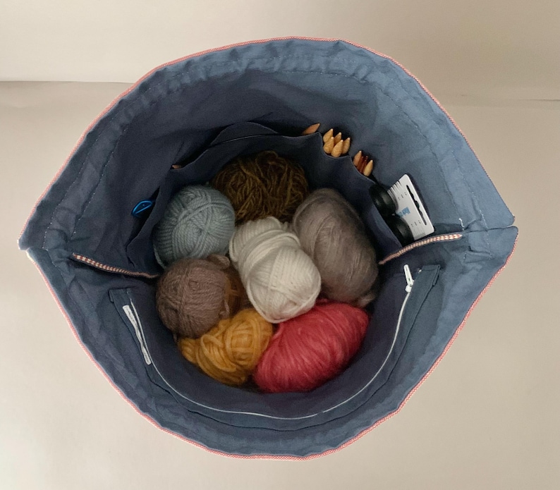 Pode incluir: Uma bolsa de projeto de tric&ocirc; de tecido azul com acabamento rosa, cheia de bolas de l&atilde; de v&aacute;rias cores, agulhas de tric&ocirc; e pequenos acess&oacute;rios. A bolsa tem v&aacute;rios bolsos e um fecho de correr.
