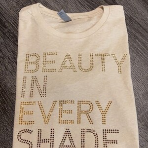 May include: A beige t-shirt with the words "BEAUTY IN EVERY SHADE" spelled out in gold rhinestones.
