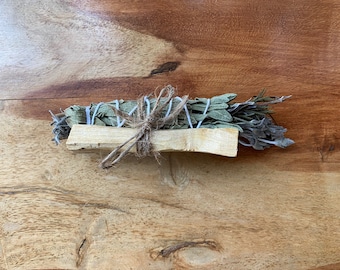May include: A bundle of dried sage and other herbs tied together with twine and white string. The bundle is sitting on a wooden surface.