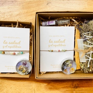 Op de afbeelding: Twee cadeaudozen met bruine deksels en witte papier vulling. Elke doos bevat een klein wit kaartje met de tekst "una ayudita para... la salud y el equilibrio" en een armband met gekleurde kralen. Een doos bevat ook een kleine kaars en een bundel gedroogde kruiden.