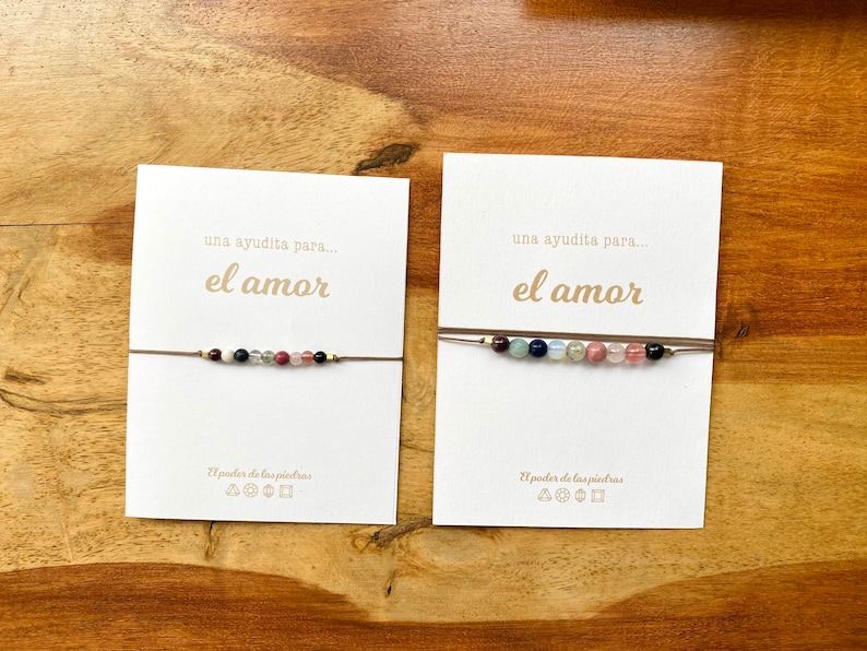Op de afbeelding: Twee kaarten met de tekst "una ayudita para... el amor" en de zin "El poder de las piedras" met symbolen. Elke kaart heeft een bruin koordarmband met gekleurde kralen.