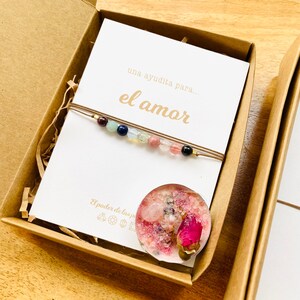 Op de afbeelding: Een bruine kartonnen doos met een witte kaart waarop "una ayuda para... el amor" staat en een kleine roze kaars met gedroogde bloemen erin. Er ligt een armband met gekleurde kralen op de kaart.