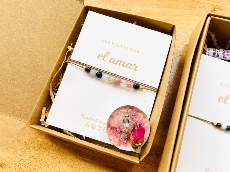 Op de afbeelding: Een bruine kartonnen doos met een witte kaart waarop "una ayuda para... el amor" staat en een kleine roze kaars met gedroogde bloemen erin. Er ligt een armband met gekleurde kralen op de kaart.