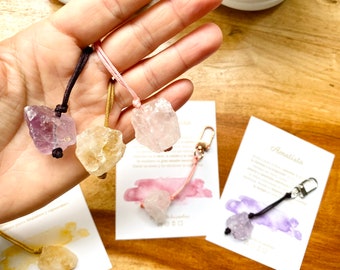 May include: Three crystal keychains with different colored stones: amethyst, rose quartz, and citrine. Each keychain has a brown cord and a gold or silver clasp. The keychains are displayed on a wooden surface with cards that have watercolor backgrounds and the names of the stones in Spanish.
