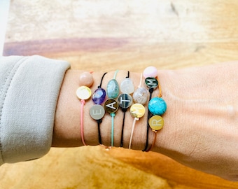 May include: A set of eight bracelets with different colored cords and gemstone beads. Each bracelet has a gold charm with a letter engraved on it. The letters spell out the word "EARTH"