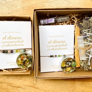 May include: Two open cardboard boxes with spiritual items. One box contains a bracelet with colorful beads and a round resin pendant with gold flakes. The other box includes a bracelet, a small bottle, a bundle of dried herbs, and a crystal.