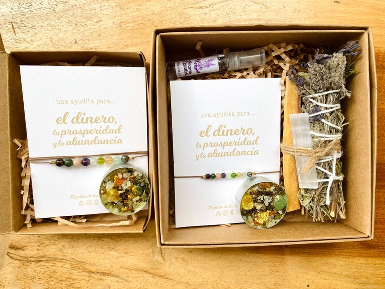 May include: Two open cardboard boxes with spiritual items. One box contains a bracelet with colorful beads and a round resin pendant with gold flakes. The other box includes a bracelet, a small bottle, a bundle of dried herbs, and a crystal.