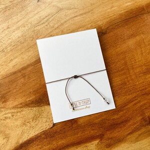 May include: A white card with a brown string tied to it, resting on a wooden surface. The card has a small logo in the bottom left corner. The string has a small, clear bead attached.