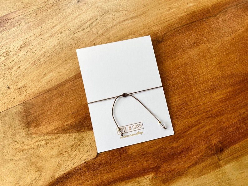 May include: A white card with a brown string tied to it, resting on a wooden surface. The card has a small logo in the bottom left corner. The string has a small, clear bead attached.