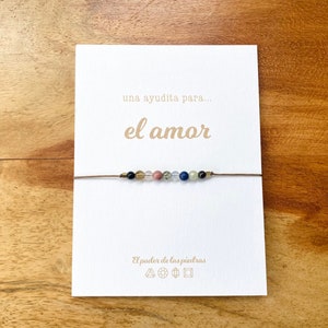 Op de afbeelding: Een witte kaart met de tekst "una ayudita para... el amor" in bruine letters. Een bruin koordarmband met gekleurde kralen is aan de kaart gebonden. De kaart heeft ook de tekst "El poder de las piedras" en symbolen.