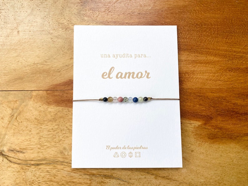 Op de afbeelding: Een witte kaart met de tekst "una ayudita para... el amor" in bruine letters. Een bruin koordarmband met gekleurde kralen is aan de kaart gebonden. De kaart heeft ook de tekst "El poder de las piedras" en symbolen.