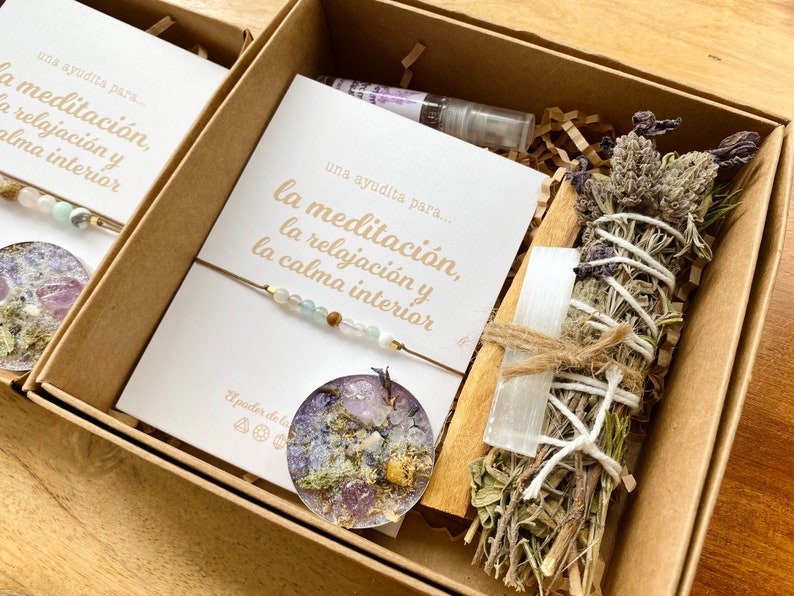Op de afbeelding: Twee cadeaudozen met een meditatiethema. Elke doos bevat een kaart met de tekst "una ayudita para... la meditaci&oacute;n, la relajaci&oacute;n y la calma interior", een armband met kralen, een klein rond harsstuk met gedroogde bloemen en een kristal, en een wierookstokje met gedroogde kruiden en een wit kristal.