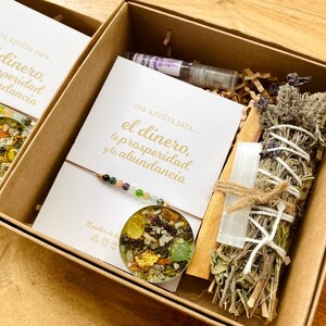 May include: Two open cardboard boxes filled with spiritual items. Each box contains a card with gold text that reads "una ayudita para... el dinero, la prosperidad y la abundancia". One box has a bracelet, a pendant, and a small bottle. The other box has a bundle of herbs, a crystal, and a wooden stick.