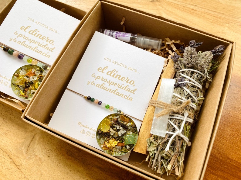 May include: Two open cardboard boxes filled with spiritual items. Each box contains a card with gold text that reads "una ayudita para... el dinero, la prosperidad y la abundancia". One box has a bracelet, a pendant, and a small bottle. The other box has a bundle of herbs, a crystal, and a wooden stick.