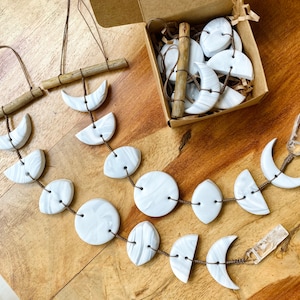 May include: Two white ceramic moon phase wall hangings with brown wooden sticks and twine. The hangings are made of different sized crescent moons and full moons.
