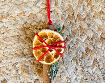May include: A handmade Christmas ornament featuring a dried orange slice, cinnamon stick, and sprig of rosemary, tied together with red twine. The ornament is suspended by a red string, perfect for holiday decor. The natural elements create a rustic, festive look.