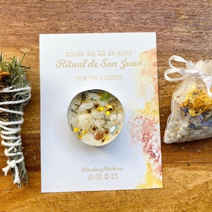 May include: An arrangement of spiritual items on a wooden surface. It includes a bundle of dried herbs wrapped in white string, a small candle with crystals and gold stars, a sachet of dried flowers, and a card with text in Spanish.