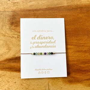 May include: A delicate bracelet featuring small, colorful gemstones strung on a thin, brown cord. The bracelet is displayed on a white card with gold text that reads "una ayudita para... el dinero, la prosperidad y la abundancia". The background is a wooden surface.