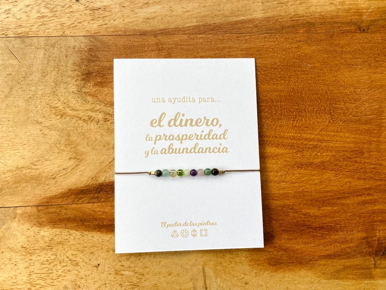 May include: A delicate bracelet featuring small, colorful gemstones strung on a thin, brown cord. The bracelet is displayed on a white card with gold text that reads "una ayudita para... el dinero, la prosperidad y la abundancia". The background is a wooden surface.