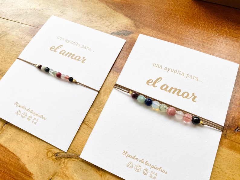 Op de afbeelding: Twee kaarten met de tekst "una ayudita para... el amor" en een bruin touwtje met gekleurde kralen. De tekst "El poder de las piedras" staat onderaan elke kaart gedrukt.