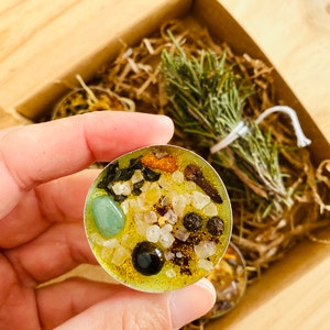 May include: A yellow tea light candle with green, black, and white crystals and herbs. The candle is in a clear glass container and is surrounded by dried herbs.