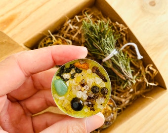 May include: A yellow tea light candle with green, black, and white crystals and herbs. The candle is in a clear glass container and is surrounded by dried herbs.