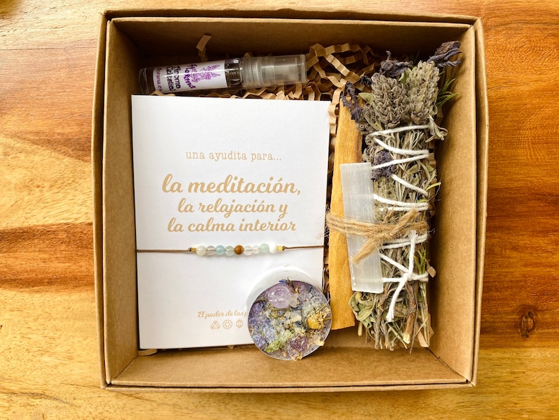 Op de afbeelding: Een bruine doos met een witte kaart met de tekst "una ayudita para... la meditaci&oacute;n, la relajaci&oacute;n y la calma interior", een klein flesje lavendelolie, een wierookstokje, een helder kristal en een armband met kralen.