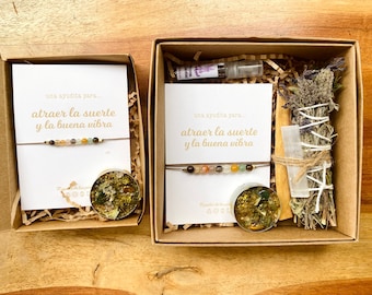 Op de afbeelding: Twee cadeaudozen met bruine papieren deksels en witte kaarteninzetstukken. De kaarteninzetstukken hebben de tekst "una ayudita para... atraer la suerte y la buena vibra" en bevatten een bruin touwtje met gekleurde kralen. Een doos bevat ook een kleine kaars met een goud-groen ontwerp en de andere doos bevat een bundel gedroogde kruiden en een klein wit kristal.