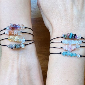 May include: Four black string bracelets with different colored gemstone chips. The bracelets are stacked on each other on a person's wrist.