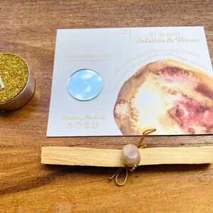 May include: A gold glitter tea light candle, a card with a circular mirror, and a bundle of Palo Santo wood tied with a stone and string. The card has text in Spanish: "21 de junio Solsticio de Verano".