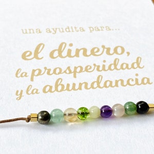 May include: A beaded bracelet with various colored gemstones, including green, purple, and clear beads, strung on a brown cord. The bracelet is displayed on a white card with gold text in Spanish.