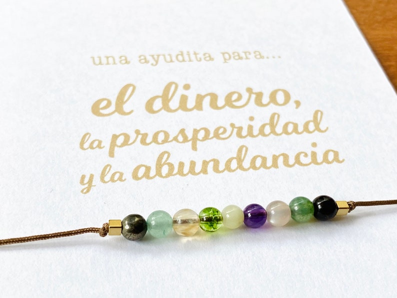 May include: A beaded bracelet with various colored gemstones, including green, purple, and clear beads, strung on a brown cord. The bracelet is displayed on a white card with gold text in Spanish.