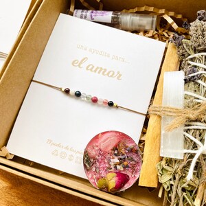 Op de afbeelding: Een cadeauset voor de liefde met een kaart waarop staat "una ayudita para... el amor" (een klein beetje hulp voor... de liefde), een armband met gekleurde kralen, een kaars met roze was en gedroogde bloemen, een wierookstokje met gedroogde kruiden en een witte kristal, en een houten stok.